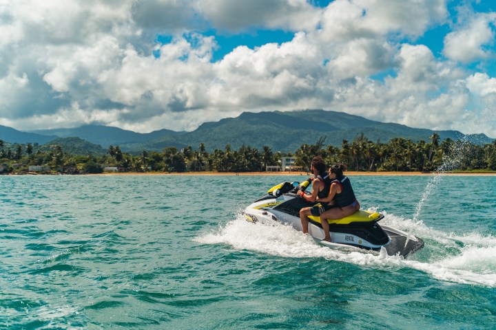 Two people riding a jet ski on tropical waters with mountains and cloudy sky.