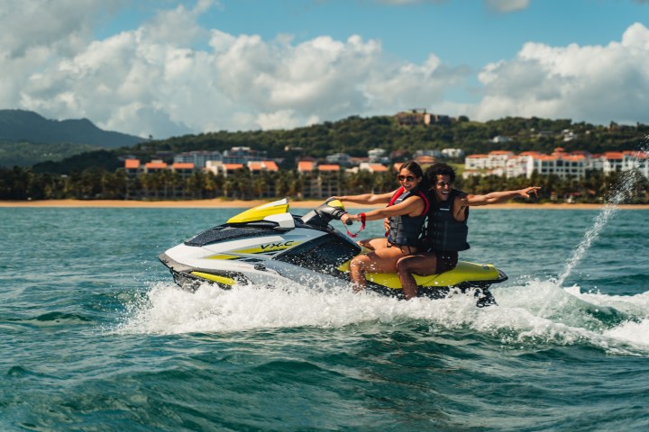 Two people riding a yellow jet ski on a sunny day with buildings and hills in the background.